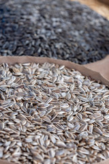 Pile of grey sunflower seeds in brown wooden bowl at food marketplace, blurred black sunflower seeds on foreground
