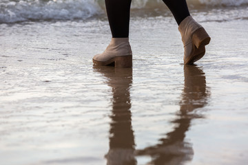 Girl's legs standing on the beach near the water line in cream-colored winter shoes and black tights, water reflection