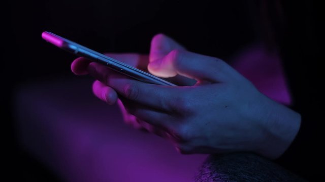 Young woman texting chatting on smart phone at night; girls finger tapping keypad, writing message on mobile devices touchscreen, blue low light; close up top side view