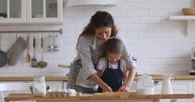 Cute funny kid daughter helping mom preparing dough for cookies together in modern kitchen interior with rolling pin, happy mother teaching little child girl learning cooking baking pastry at home