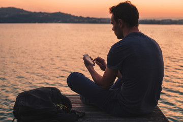 man using mobile internet on smartphone while sitting outside near the lake at sunset