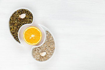 Pumpkin and sunflower seeds lie on a white background with garlic cloves in the form of a Yin-Yang sign, with an orange cut between them. Concept of healthy food, organic food. The view from the top.