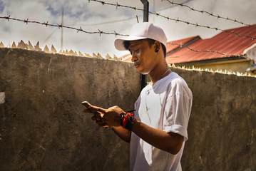 Brown-skinned Indonesian boy dressed entirely in white with a cap, watch, and bracelets next to a gray cement wall with glass fences writing a text message with his smartphone. Face profile side. Asia
