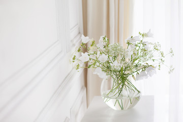 Bouquet of gentle bells in vase. Morning light in the room. Soft home decor, glass vase with white flowers on  white wall background and on wooden table. Interior. Greeting card. Copy space.