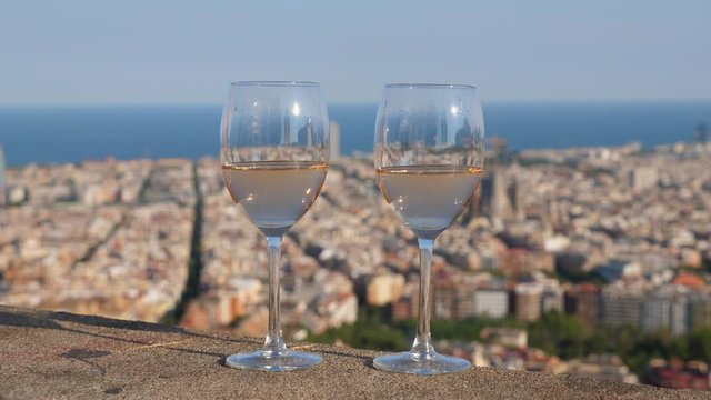 Two glasses of light wine stand at parapet, blurred Barcelona cityscape on background, Mediterranean sea on horizon. Sunny evening time, still life shot at outdoors