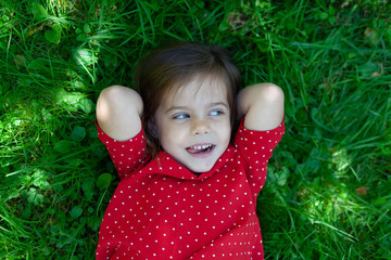 Top view of beautiful little girl is lying on a green grass, smiling and looking at the camera