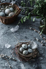 Modern Easter eggs in wicker basket on blue background with nest, feathers and spring flowers. Happy Easter holiday