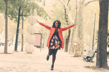 Cheerful woman with raised hands walking in park. Beautiful excited young African American woman walking on street and smiling at camera. Happiness concept