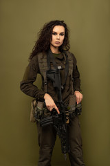 Military woman in green uniform with a vest unloading with a machine gun in her hands on a khaki background.