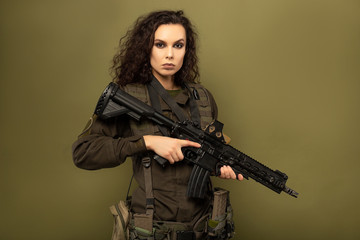 Fototapeta premium Military woman in green uniform with a vest unloading with a machine gun in her hands on a khaki background.