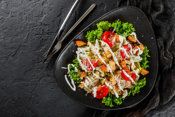 Healthy Caesar salad of fresh vegetables with tomatoes, lettuce, croutons, grilled chicken breast, cheese parmesan and sauce in plate on black stone background. Healthy food, top view