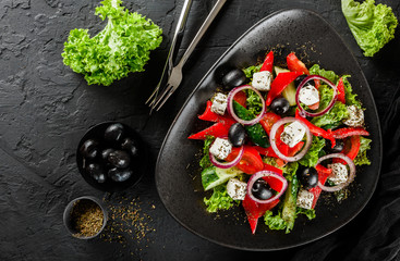Healthy greek salad of fresh vegetables with tomatoes, lettuce, bell pepper, cucumbers, olives, feta cheese in plate on black stone background. Healthy food, vegetarian dieting, top view