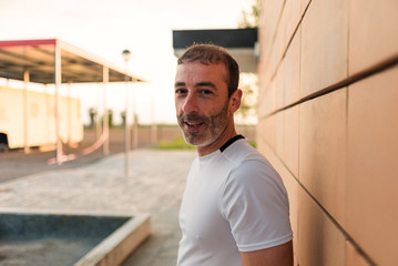 Caucasian athlete man in white t-shirt, next to an orange brick wall, looking happy at the camera.