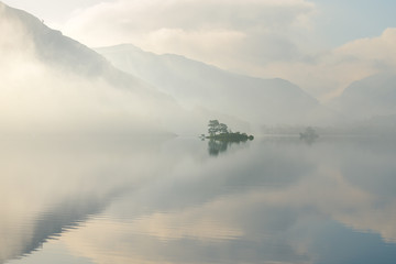 Obraz na płótnie Canvas Isolated Island Reflected In A Calm Still Lake On A Peaceful Misty Summer Morning. Lake District, UK.