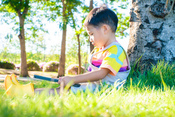 Adorable boy sitting under tree in city park use tablet computer on green grass