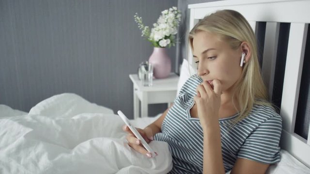 Happy Beautiful Young Woman with wireless headphones lying on the Bed and using smartphone