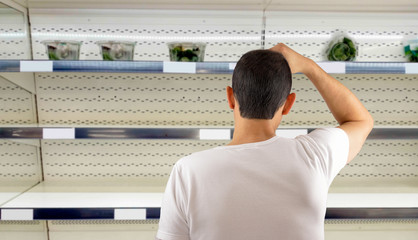 Rear view of a man at empty shelves in shop scratches in nape.Crisis concept