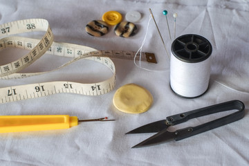 Needle and pin on white thread rolls for hand sewing on white cloth with tape measure and sketch yellow chalk with scissors cut and seam ripper on the fabric close-up.