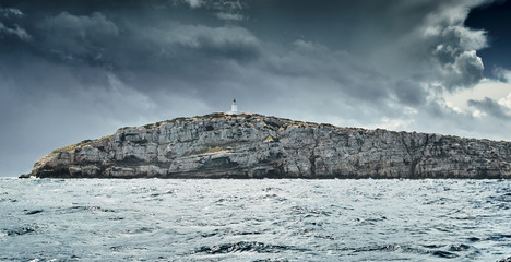 Lighthouse is on edge of the rock, the landscape of the balearic sea and improbable mountains, azure water, the storm sky, mountains