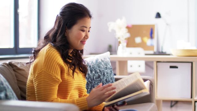 leisure, literature and people concept - young asian woman reading book and drinking water at home