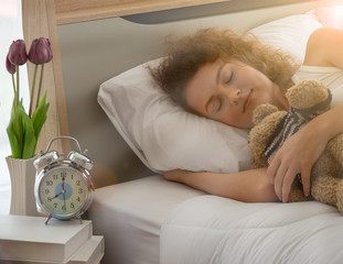 Beautiful young lady is sleeping on the bed with alarm clock and purple tulip on the side table.