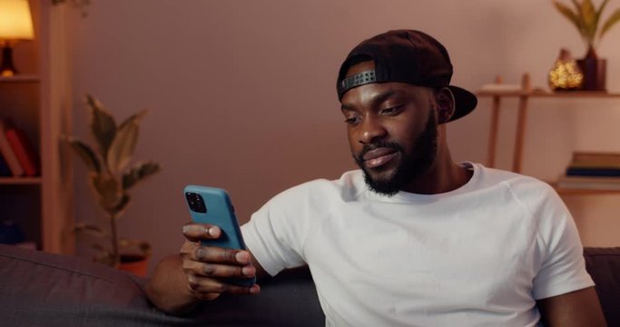 Close up of african guy using his smartphone while sitting on sofa at home. Bearded man wearing cap scrolling news feed and looking at mobilephone screen while spending free time.