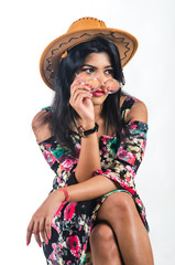 Young woman in a floral pattern dress wearing a cowboy hat and sitting cross legged turning to her left and perching a colorful glasses on her nose with her right hand and looking at a distance