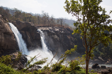 Fototapeta na wymiar A huge waterfall Athirapally in Kerala, India. White streams of water of the waterfall break into small splashes and flow in a strong stream, Indian tourists look at the attraction