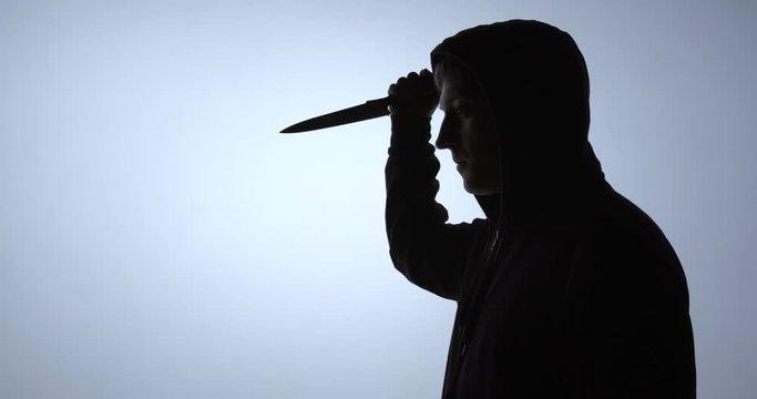 Silhouette of a criminal in the hood raises a knife up and makes a blow
