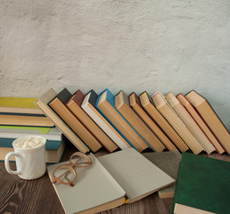 Time for yourself, the pleasure of reading. A row of books, an open book, glasses, a Cup of cocoa with marshmallows against a gray concrete wall. Free space.