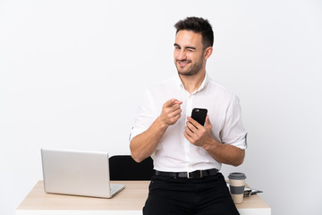 Young business man with a mobile phone in a workplace points finger at you