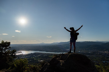 Silhouette of girl in hat, stains on a rock and spreads her hands to the sides. A city, mountains, sea and sun at the background
