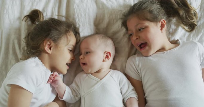 Authentic close up shot of happy little sisters are cuddling a newborn brother just woke up in a bed with white sheet in a morning.Concept: children,family, care,love, healthcare, comfort,authenticity