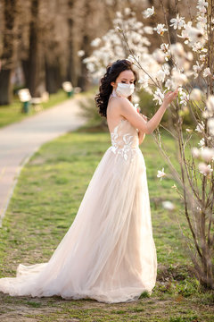 Beautiful young bride in a wedding dress and a white medical mask on her face near a blooming magnolia. Crown virus. Kovid-19 protects.