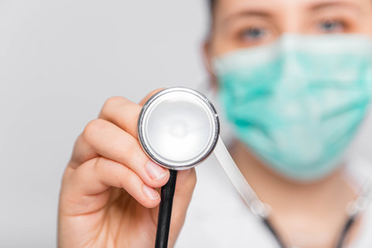 Focus On The Acoustic Head Of A Stethoscope In The Hands Of A Young Woman In A White Coat And Medical Mask. The Nurse Listens To The Heartbeat.