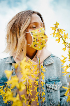 Attractive young woman in yellow medical mask standing in the yellow blooming tree in the sunny day. Natural light