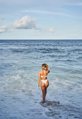 Slim woman in bikini carrying surfboard and walking on wet sand towards waving sea on beach