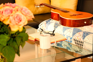 Book, glasses, roses and a mug with tea on modern glass table. An acoustic Spanish guitar is lying on an old rug on a black leather sofa. Living room interior home decor concept.