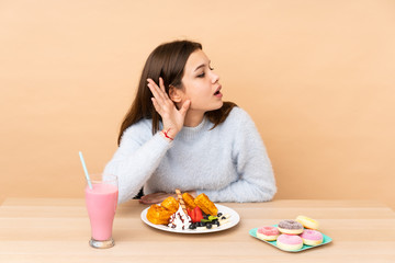 Teenager girl eating waffles isolated on beige background listening to something by putting hand on the ear