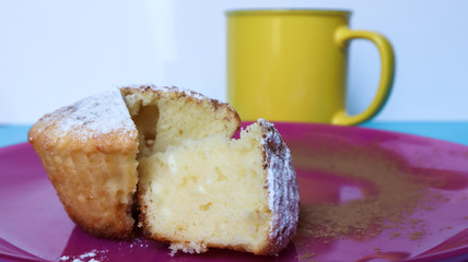 Side view of one cottage cheese sprinkled with icing sugar on a pink plate next to a yellow cup of coffee or tea. Dessert, a small cupcake. white baked cookies with an airy texture. Copy space