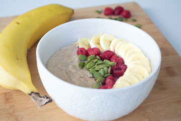 Homemade healthy breakfast: oatmeal porridge with banana, ripe raspberries and pumpkin seeds with ingredients in the background