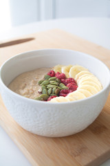 Homemade healthy breakfast: oatmeal porridge with banana, ripe raspberries and pumpkin seeds with ingredients in the background