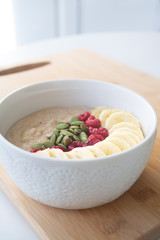 Homemade healthy breakfast: oatmeal porridge with banana, ripe raspberries and pumpkin seeds with ingredients in the background