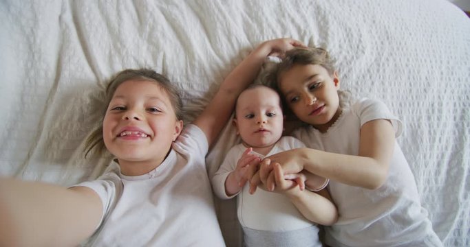 Authentic shot of happy little sisters with newborn brother are making a selfie or video call to parent or relatives in a bed. Concept of technology, new generation,family, connection, childhood, auth