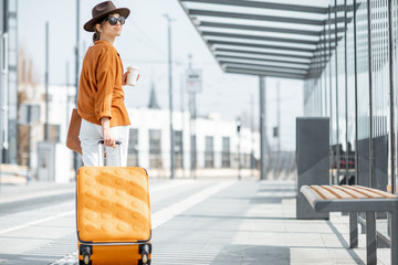 Young female traveler walking with a yellow suitcase at the modern transport stop outdoors, back...