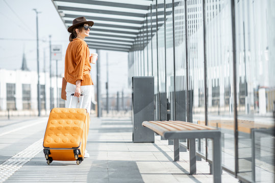 Young female traveler walking with a yellow suitcase at the modern transport stop outdoors, back view. Concept of an urban transportation and travel