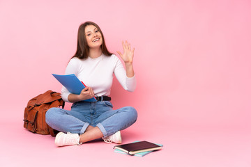Teenager caucasian student girl sitting on the floor isolated on pink background saluting with hand with happy expression