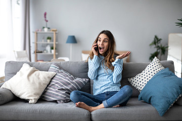 Obraz premium Woman emotionally talking on a mobile while sitting at home on the couch. Portrait of a positive girl.