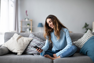 Obraz premium Online chat. Smiling woman talking on smartphone with friends while sitting on sofa at home.