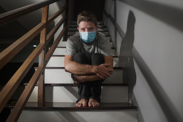 dark portrait of young scared and worried man in protective mask sitting on stairs at home...